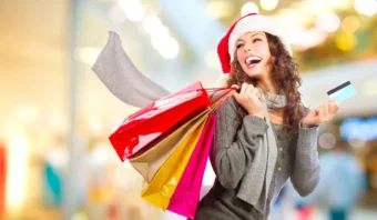 Una mujer que lleva un gorro y una bufanda de Papá Noel sostiene varias bolsas de compras y una tarjeta de crédito, sonriendo en medio del bullicio de las compras de fin de año en un entorno interior muy iluminado.