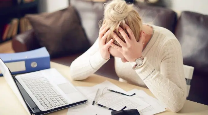 Una persona se sienta en una mesa con una computadora portátil y papeles, sosteniendo su cabeza en señal de frustración, en medio de una crisis inminente.