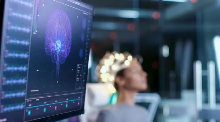 Una persona está sentada con un dispositivo de monitoreo cerebral, mientras la pantalla de computadora frente a ella muestra una imagen digital del cerebro, que refleja las maravillas de las neurociencias aplicadas.