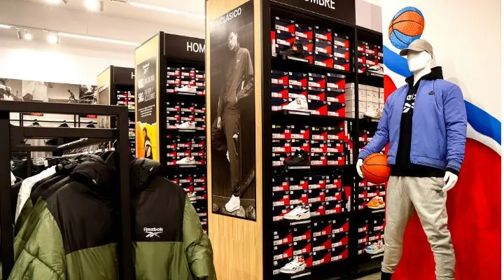 Un maniquí con ropa deportiva sostiene una pelota de baloncesto en la sección Reebok de Outlet Puebla Premier. Los estantes están repletos de cajas de zapatos, cerca se exhiben chaquetas y un gran póster de alguien con ropa deportiva adorna la pared.