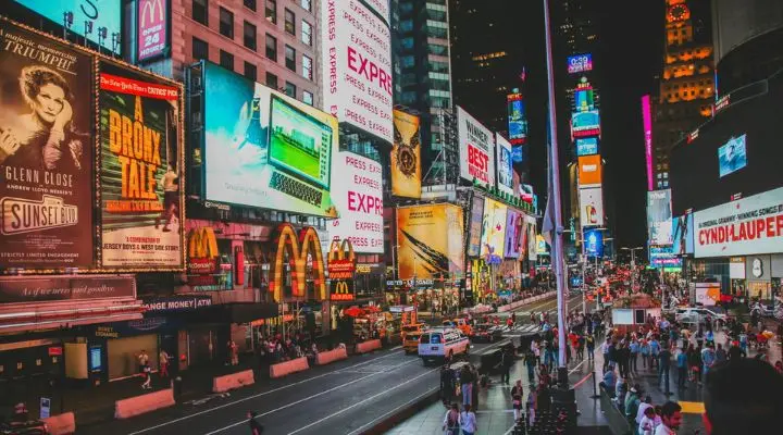 Times Square por la noche estalla con una saturación publicitaria, sus brillantes anuncios y vallas publicitarias iluminan el cielo. La gente camina por la calle, reuniéndose en este bullicioso centro de intensa energía.