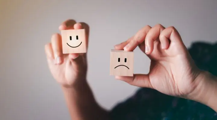 Dos manos sostienen bloques de madera, una con una cara sonriente y la otra con una cara triste, sobre un fondo neutro. Esta sencilla escena refleja sutilmente el equilibrio que buscamos en materia de sostenibilidad: entre alegría y preocupación, progreso y conservación.