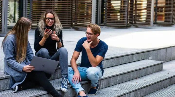 Tres personas están sentadas en unas escaleras al aire libre, enfrascadas en una animada conversación. Una sostiene un teléfono, otra una computadora portátil, equipada con herramientas de gamificación que despiertan la curiosidad. La tercera persona, sonriendo alegremente, parece deleitarse con el dinámico intercambio de ideas.