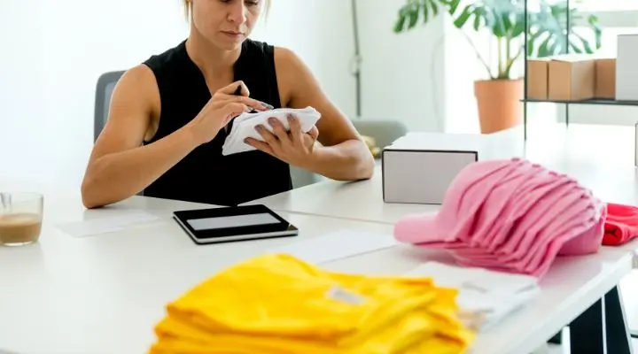 Una persona revisa prendas de vestir en una mesa, rodeada de pilas de camisetas amarillas y rosas dobladas, con una tableta y una caja cerca de merch.