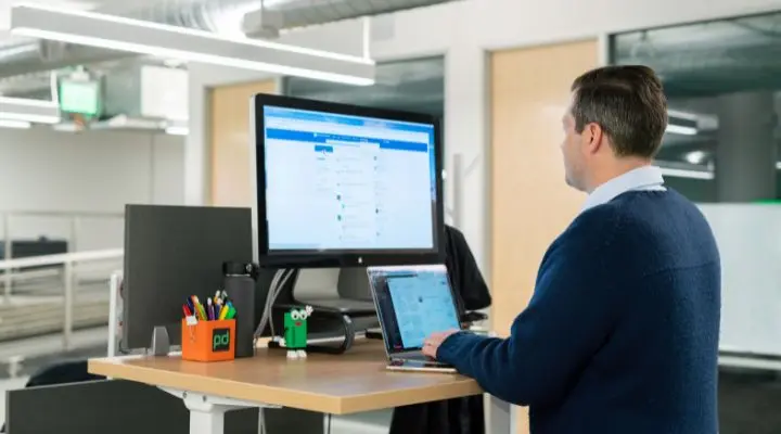 Una persona trabaja con una computadora portátil en un escritorio de pie, con un monitor grande que muestra su feed de LinkedIn.