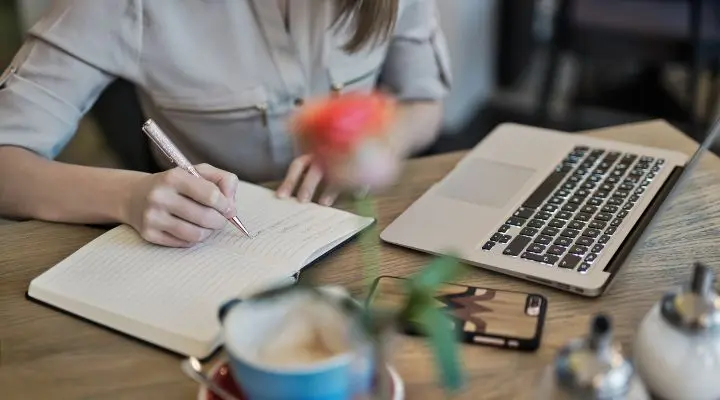 Una persona toma notas meticulosamente en un cuaderno sentado a la mesa, rodeada de un ordenador portátil y un teléfono, mientras se concentra en la comercialización de marketing de contenidos. En primer plano, una flor suavemente difuminada añade un toque artístico a la escena.