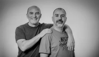 Dos hombres posando juntos, uno con el brazo sobre el hombro del otro. Ambos llevan camisetas informales. La imagen es en blanco y negro.