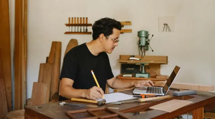 En una muestra de la resiliencia, una persona con una camisa negra trabaja diligentemente con su computadora portátil en un taller de carpintería, rodeada de una variedad de herramientas y materiales de madera.