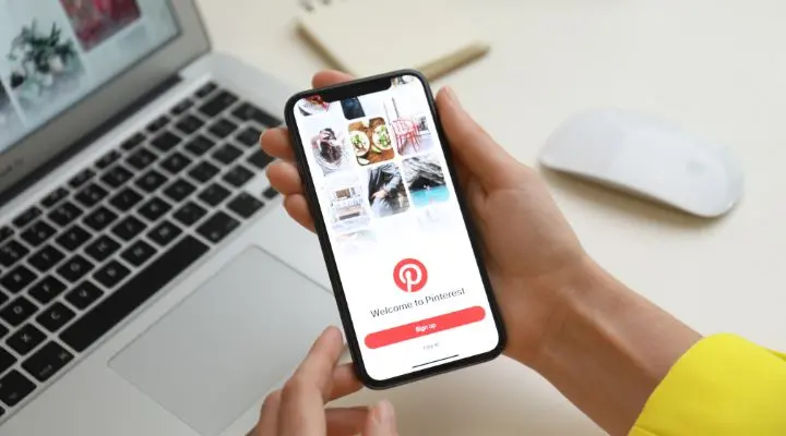 Una persona sostiene un teléfono inteligente que muestra la colorida pantalla de bienvenida de la aplicación Pinterest, con el telón de fondo de una elegante computadora portátil y un mouse inalámbrico en el escritorio, listo para la inspiración creativa.