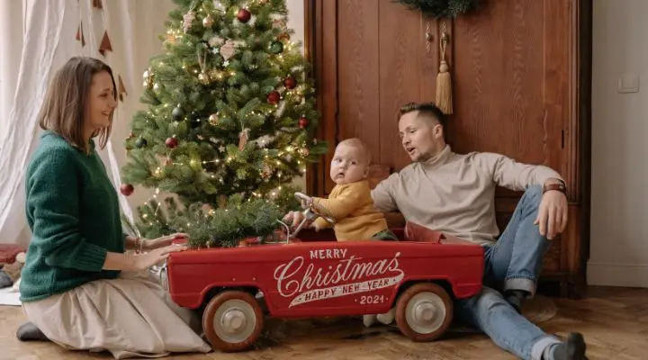 Una familia se sienta junto a un árbol de Navidad bellamente decorado y acoge el espíritu navideño. Un niño viaja feliz en un carro rojo adornado con la frase "Feliz Navidad y próspero año nuevo 2021", capturando un momento que combina alegría festiva y un sutil atractivo del neuromarketing navideño.