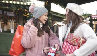 Dos mujeres vestidas con ropa de invierno sonríen y sostienen bolsas de compras y regalos envueltos en un mercado al aire libre.