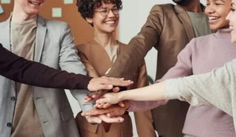 Un grupo de personas vestidas de negocios se encuentran paradas en un círculo, sonriendo y juntando sus manos en una muestra de trabajo en equipo y cooperación de las herramientas digitales.