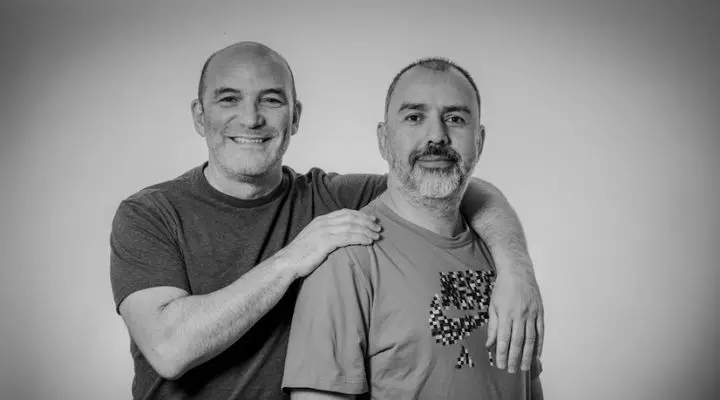 Dos hombres posan juntos en una foto en blanco y negro, irradiando camaradería. Uno sonríe con su mano sobre el hombro del otro, mostrando su vínculo. Ambos visten camisetas informales que insinúan un ambiente relajado de Bombay agencia de comunicación, como si promocionaran la última campaña de una agencia de comunicación con un encanto natural.