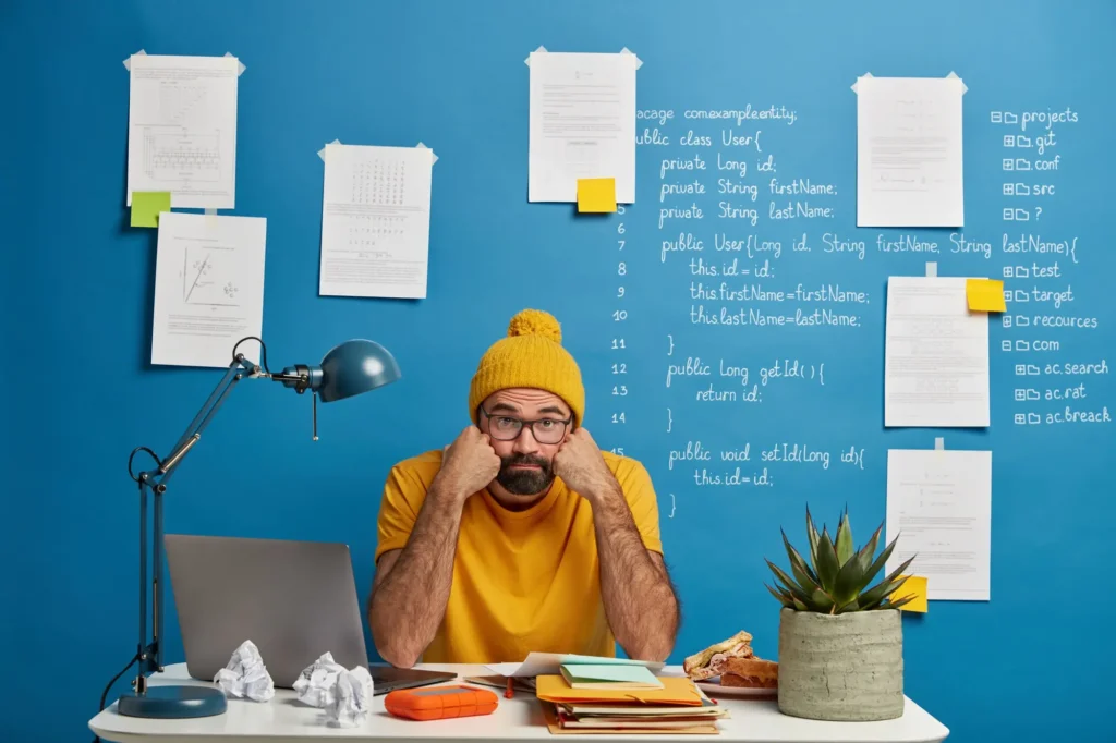 Una persona con una camiseta amarilla y un gorro está sentada en un escritorio desordenado y parece frustrada por la postergación de tareas. y procrastinación. Detrás hay una pared azul llena de códigos y papeles dispersos.