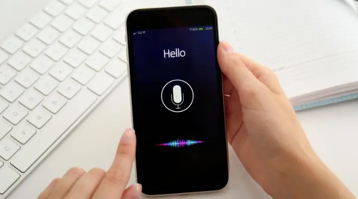 Una persona sostiene un teléfono inteligente que muestra un asistente de voz con la palabra "Hola" en la pantalla, lo que resalta la importancia de la optimización de la búsqueda por voz o Voice Search Optimization . En el fondo, hay un teclado y una computadora portátil, lo que crea una combinación perfecta de tecnología y herramientas tradicionales.