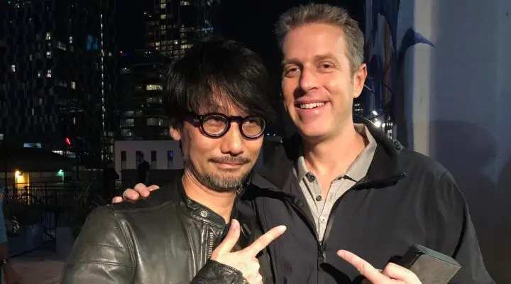 Dos hombres están de pie juntos al aire libre, de noche, sonriendo y señalándose el uno al otro. Hideo Kojima gafas y una chaqueta de cuero; Geoff Keighley sostiene un teléfono. Al fondo se ven edificios de la ciudad.