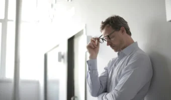 Un hombre con una camisa clara, que encarna la resiliencia, sostiene sus gafas mientras se apoya contra una pared en una habitación luminosa.