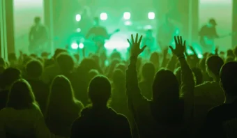 Una multitud disfruta de una actuación de música en vivo en un lugar iluminado con luz verde, con siluetas en el escenario y un miembro de la audiencia levantando las manos.