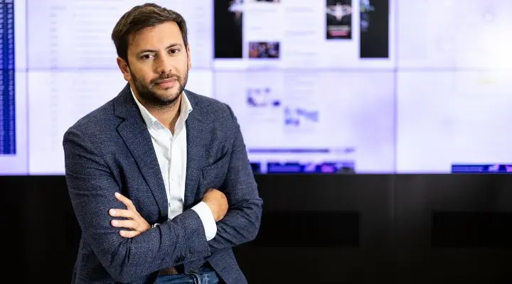 Un hombre con una chaqueta azul y una camisa blanca se sienta con los brazos cruzados frente a una pantalla que muestra diversos contenidos digitales, mientras se prepara para discutir las ideas de la Junta de Asesores sobre IA.