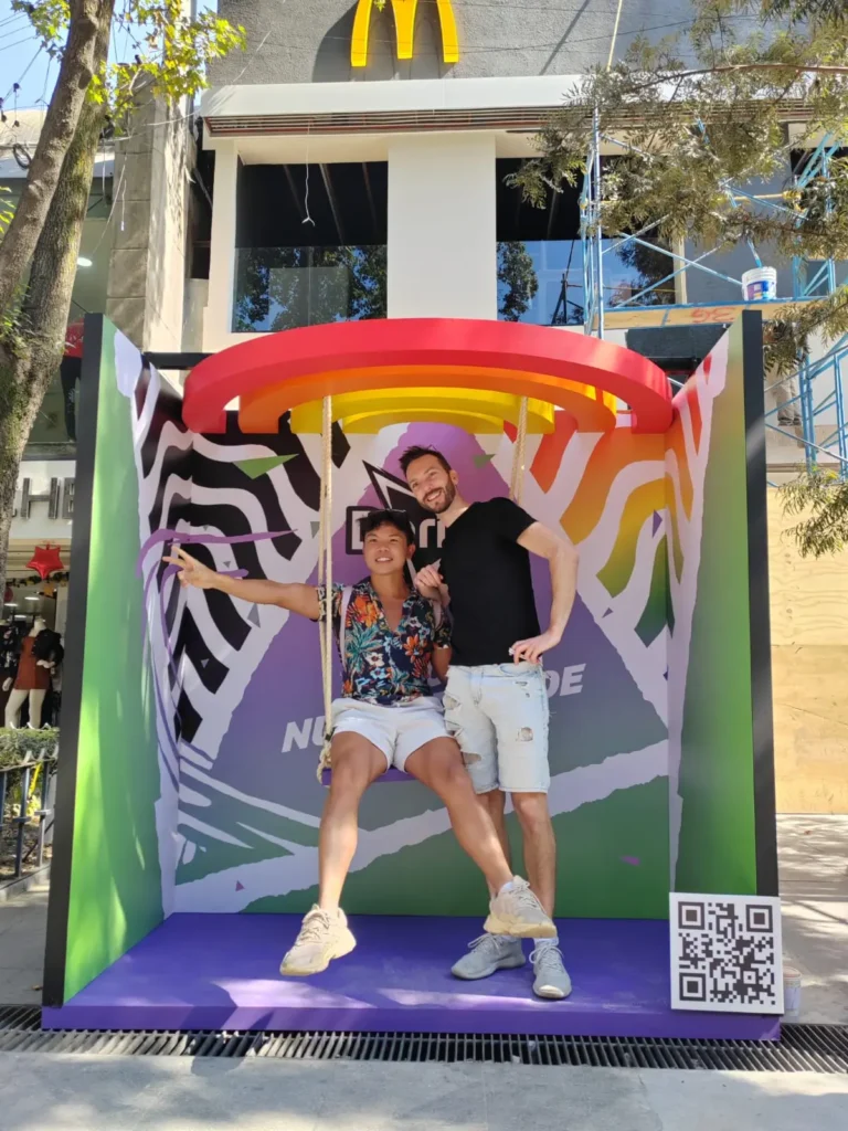 Dos personas posan en un columpio colorido con un código QR y un arco de McDonald's en el fondo, resaltando su apoyo a The Trevor Project.