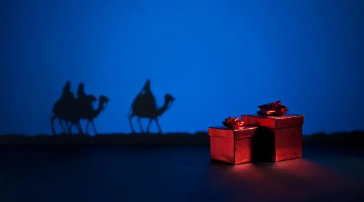 Tres figuras oscuras sobre camellos, que recuerdan a los Queridos Reyes Magos, se acercan a tres cajas de regalo rojas, sobre un fondo azul.