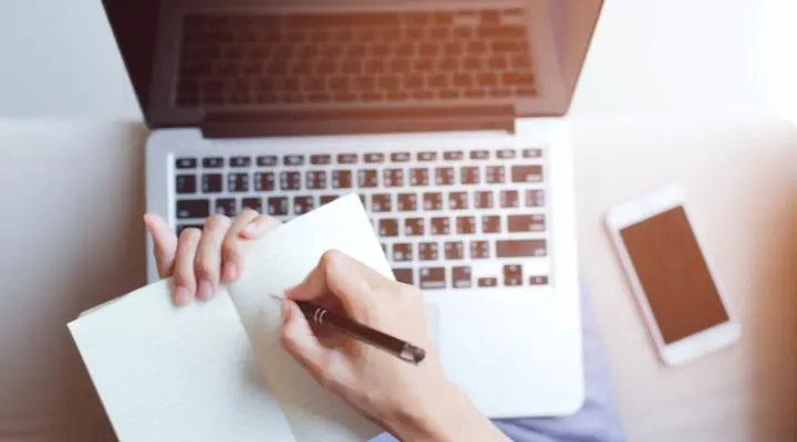 Persona concentrada en la redacción de textos, tomando notas en un cuaderno con un bolígrafo. Cerca, una computadora portátil abierta y un teléfono inteligente mejoran el espacio de trabajo productivo.