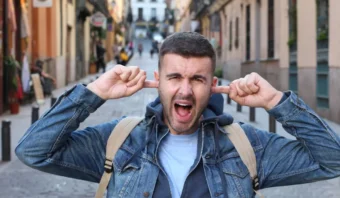 Un hombre parado en la calle se tapa los oídos con los dedos, tratando desesperadamente de romper el ruido, pareciendo gritar o reaccionar ante la cacofonía abrumadora.