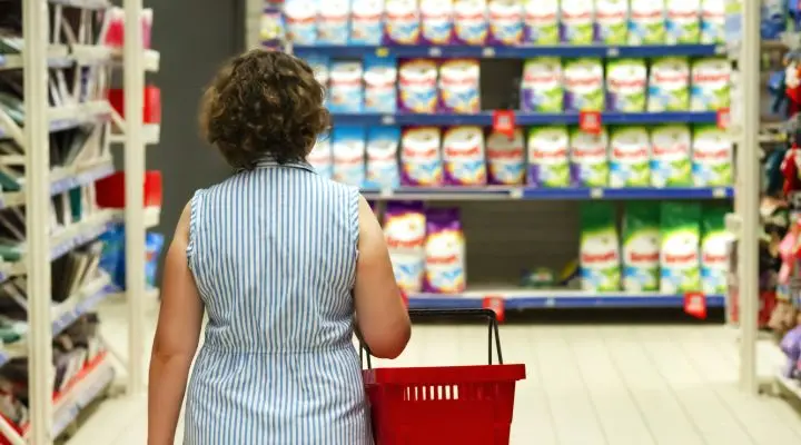 Una persona con una cesta de la compra se detiene frente a una segmentación con diversos detergentes para ropa, considerando atentamente sus opciones en la tienda.