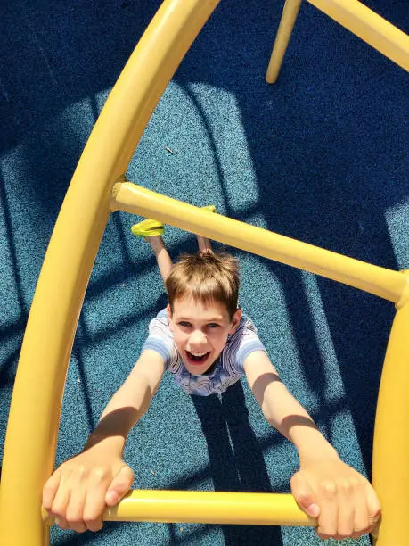 Un niño sonriente sube una escalera amarilla de juegos infantiles en un parque infantil, visto desde arriba, con una superficie de goma azul debajo.