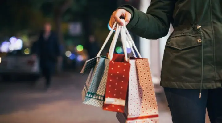 Una persona con una chaqueta oscura, envuelta en la emoción de las compras navideñas, sostiene varias bolsas de compras estampadas en una calle poco iluminada.