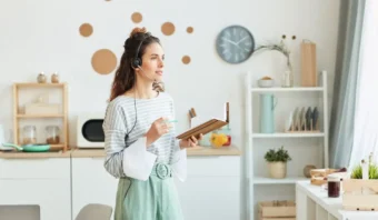 En la cocina, una mujer con auriculares está de pie, pensativa, junto a la ventana, sosteniendo un cuaderno y un bolígrafo. Mientras mira hacia afuera, su concentración en el desarrollo de su marca personal es palpable en este tranquilo momento de reflexión.