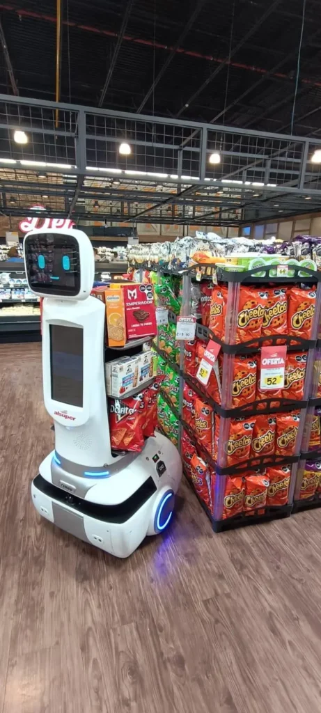 Un Shopperbot está ubicado en el pasillo de un supermercado, entre estantes repletos de diversos bocadillos, incluidas bolsas de Cheetos y galletas saladas.