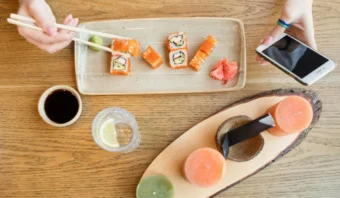 Una persona usa palillos para tomar sushi de un plato rectangular. Cerca hay salsa de soja, un vaso de agua con limón, una vela redonda y un teléfono inteligente sobre una mesa de madera.