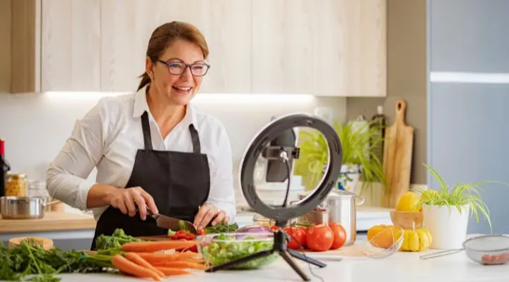 Una mujer con gafas y delantal sonríe mientras pica verduras; sus movimientos quedan perfectamente captados por un teléfono inteligente en threads colocado sobre un trípode. A través de este vídeo, entrelaza sin esfuerzo los hilos del arte culinario y la creatividad alegre.