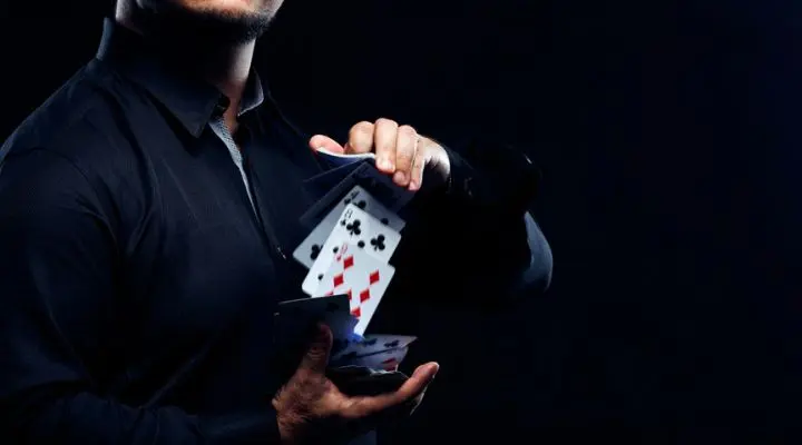 Una persona con una camisa oscura baraja hábilmente una baraja de cartas, mostrando varias cartas desplegadas en el aire sobre un fondo oscuro, que recuerda a una elegante campaña de publicidad.