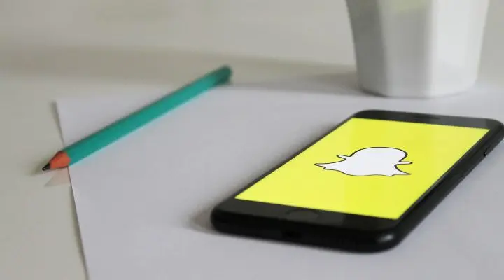 Un teléfono inteligente que muestra el logotipo de Snapchat reposa elegantemente sobre una hoja de papel blanco al lado de un lápiz verde azulado, adyacente a una taza, insinuando sutilmente el mundo digital lleno de matices que gigantes como Meta están dando forma.