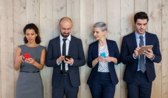 Cuatro personas vestidas de negocios están paradas frente a una pared de madera, cada una usando un teléfono inteligente o una tableta.