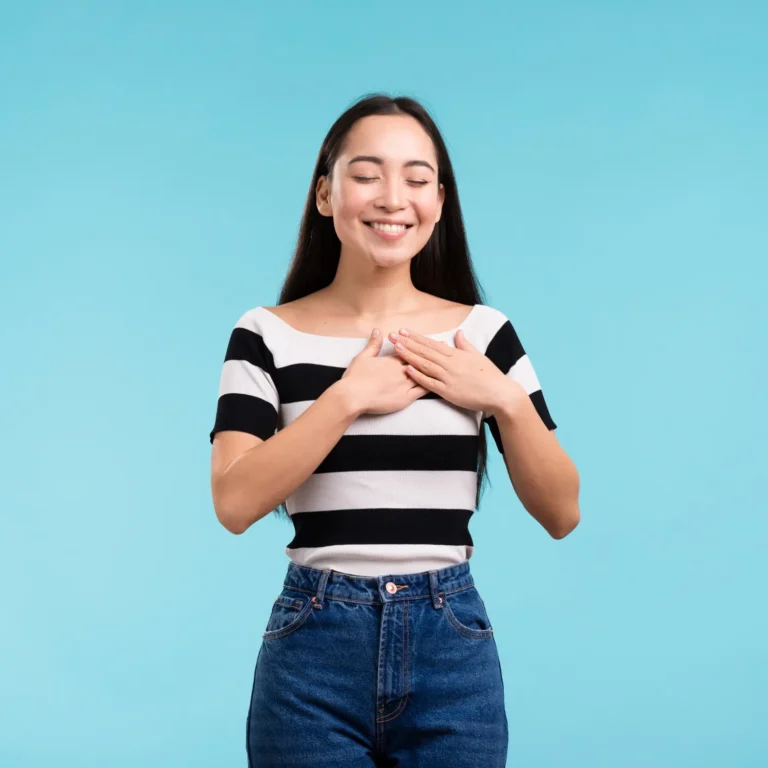 Una persona con cabello largo, vistiendo una camisa a rayas y jeans, está parada sobre un fondo azul, con las manos en el pecho, exudando una sensación de autoestima mientras sonríe con los ojos cerrados.