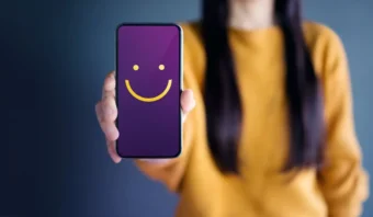 Persona con un suéter amarillo sosteniendo un teléfono inteligente con una pantalla violeta que muestra una cara sonriente amarilla.