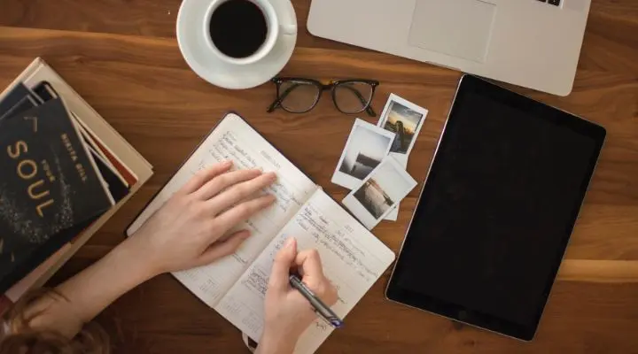 Una persona escribe en un cuaderno contenido sobre una mesa de madera con una computadora portátil, una taza de café, vasos y fotos cerca.