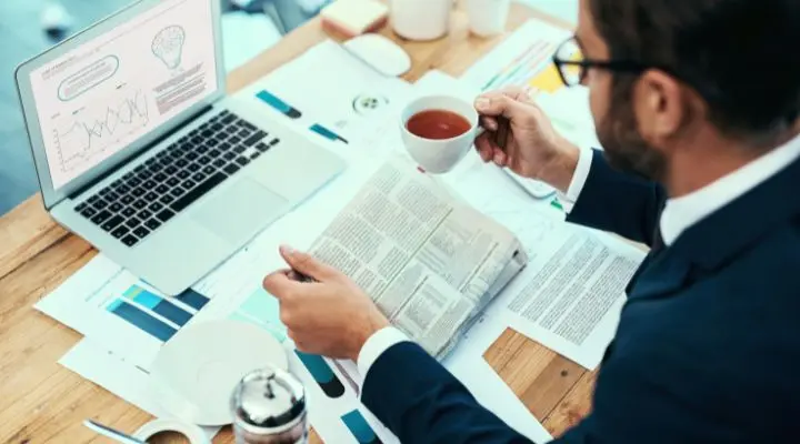 Una persona con traje lee un periódico sobre relaciones públicas mientras sostiene una taza de té en un escritorio con una computadora portátil que muestra gráficos y documentos.