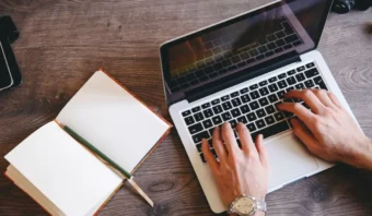 Una persona crea contenido mientras escribe en un teclado portátil al lado de un cuaderno abierto con un lápiz, todo sobre un escritorio de madera.