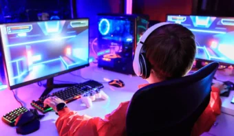 Persona que usa auriculares mientras juega un videojuego en una computadora con luces de colores, usando un teclado y un mouse, sentada en un escritorio en una habitación con poca luz.