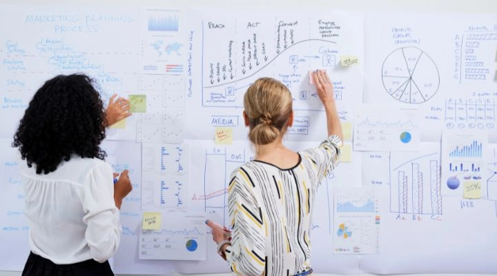 Dos mujeres están paradas frente a una pizarra cubierta de gráficos, cuadros y notas, discutiendo estrategias de arquitectura comercial y agregando notas adhesivas al tablero.