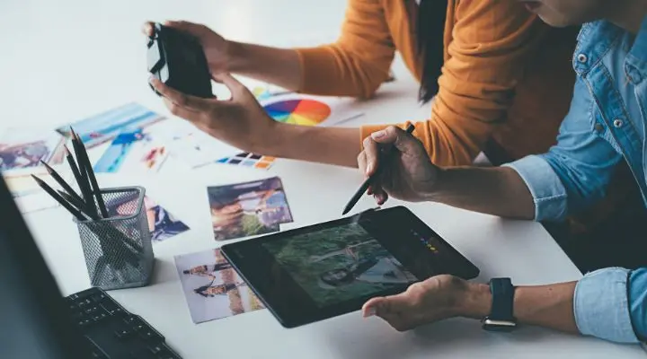 Dos personas trabajan en un escritorio, concentrándose en crear su marca personal con una tableta y una cámara. Una usa un lápiz en la tableta mientras la otra sostiene la cámara con destreza. Fotos, una rueda de colores y bolígrafos están esparcidos por el escritorio, creando el escenario para una marca creativa.