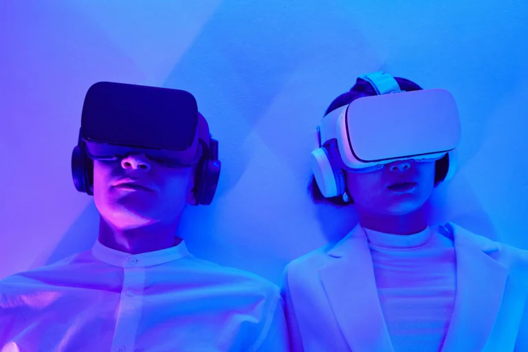 Dos personas que usan cascos y auriculares de realidad virtual se sientan contra una pared, iluminada por luces azules y violetas, creando una escena atractiva, perfecta para el marketing experiencial.