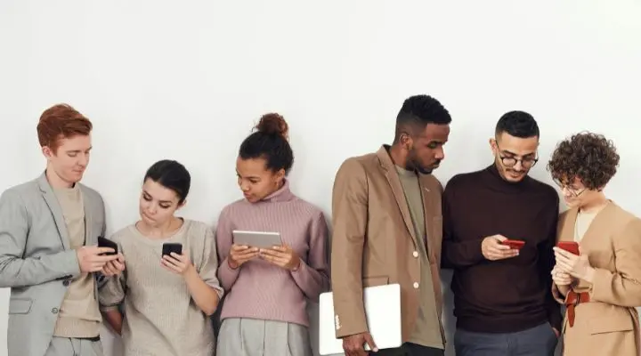 Un grupo de seis personas están de pie frente a una pared blanca, vestidas informalmente y profundamente concentradas en sus dispositivos, probablemente explorando las últimas tendencias en marketing móvil.