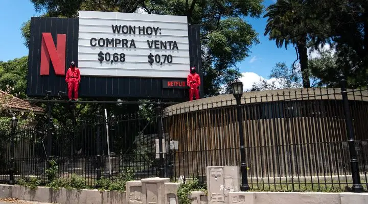 Un cartel dice "Won Hoy: Compra Venta $0.68 $0.70" junto a un enorme logotipo de Netflix. Dos personas con trajes rojos están de pie encima, enmarcadas por árboles y vallas, lo que recuerda a una escena creada por Febrero MADE.