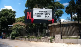 Cartelera de Netflix con dos maniquíes con monos rojos sentados sobre ella. El cartel muestra el tipo de cambio: "Won Hoy: Compra $0,68, Venta $0,70". Rodeado de árboles y una valla.