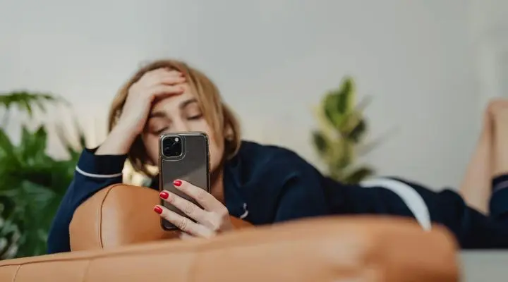 Una persona descansando en un sofá mira su teléfono inteligente, disfrutando del tiempo en redes sociales, mientras apoya la cabeza en una mano.